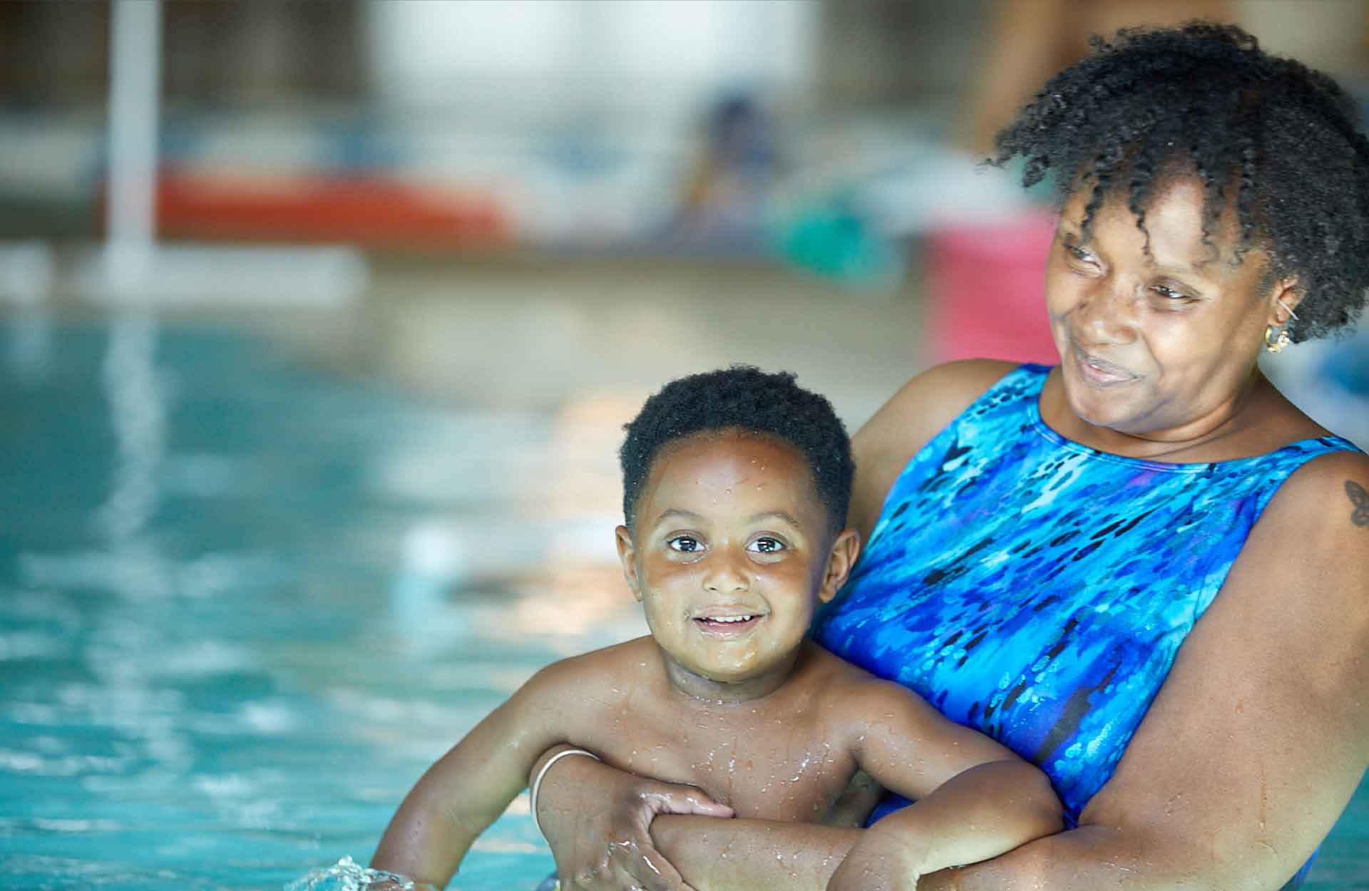 Black mom with a blue bathing suit holder her black son in the water while he is smiling at the camera