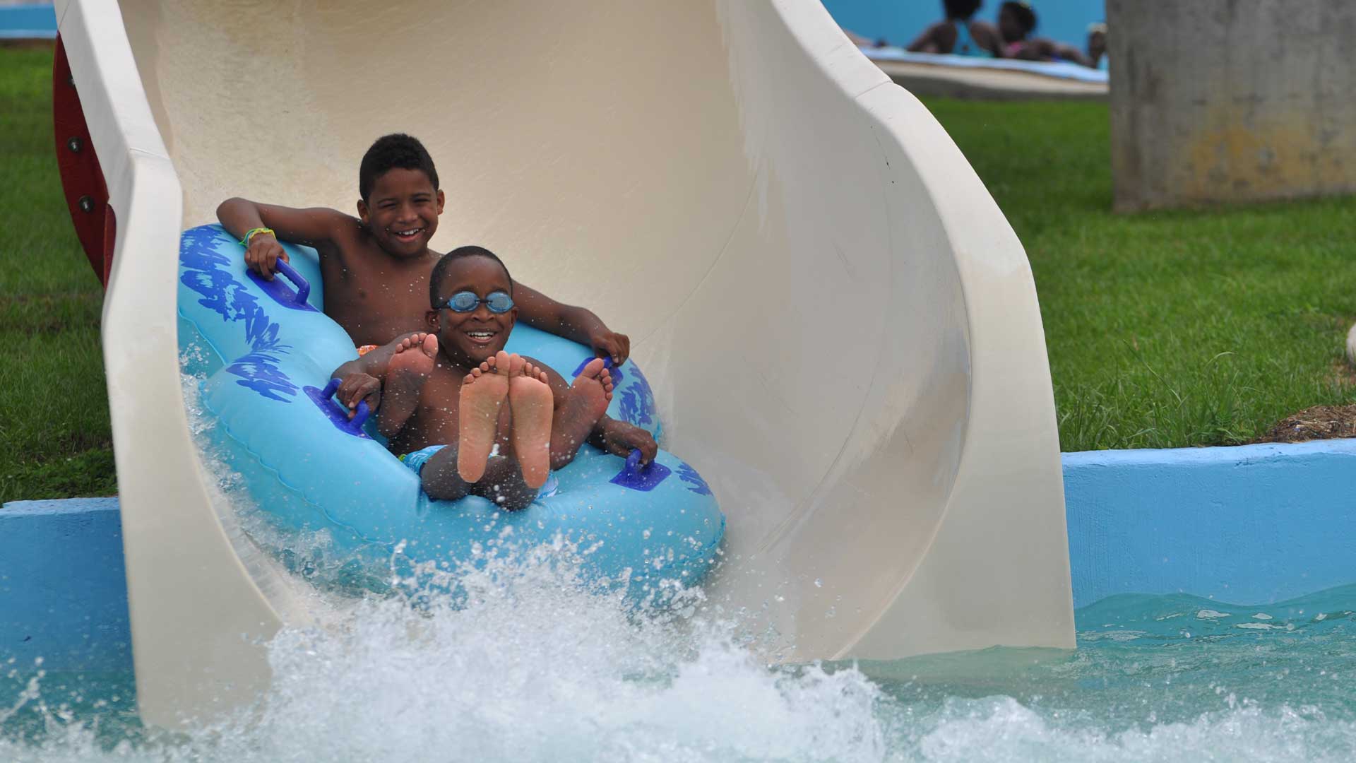 two young black boys on a float coming down a waterslide into the pool water