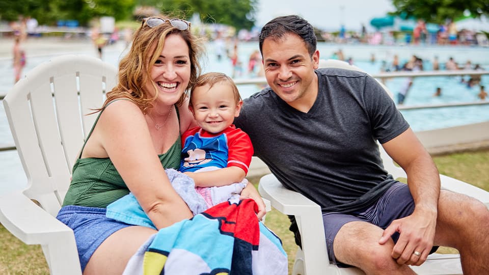 Young family, toddler in Mother's lap sitting in the grass next to the Wave Pool