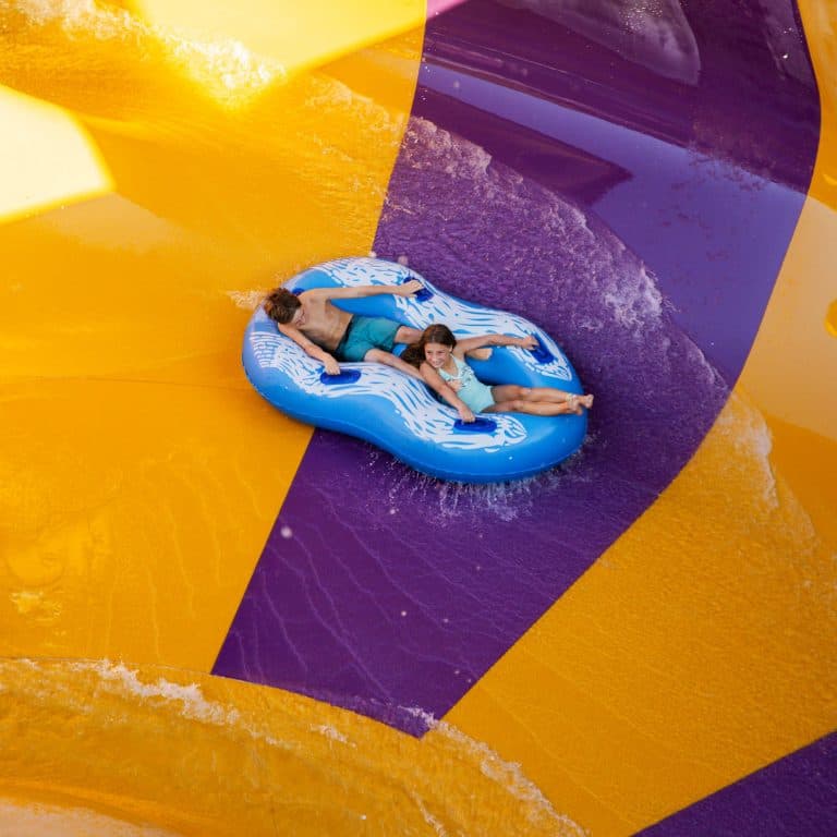 Two people ride a blue inflatable tube down a vibrant yellow and purple water slide. They appear excited and are holding on tight as water splashes around them.